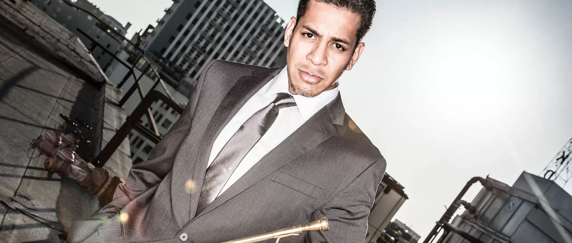 Barry Perkins in a gray suit and tie holding his trumpet on a rooftop