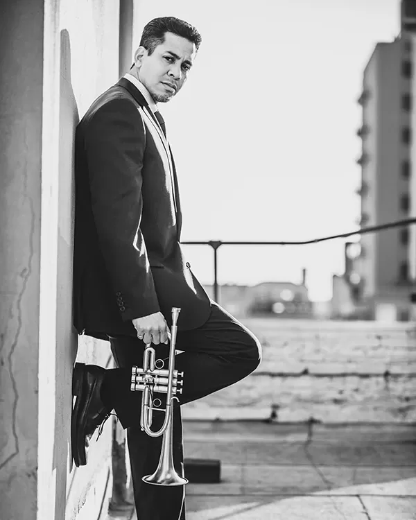Barry Perkins in a gray suite holding his trumpet in front of a city scene