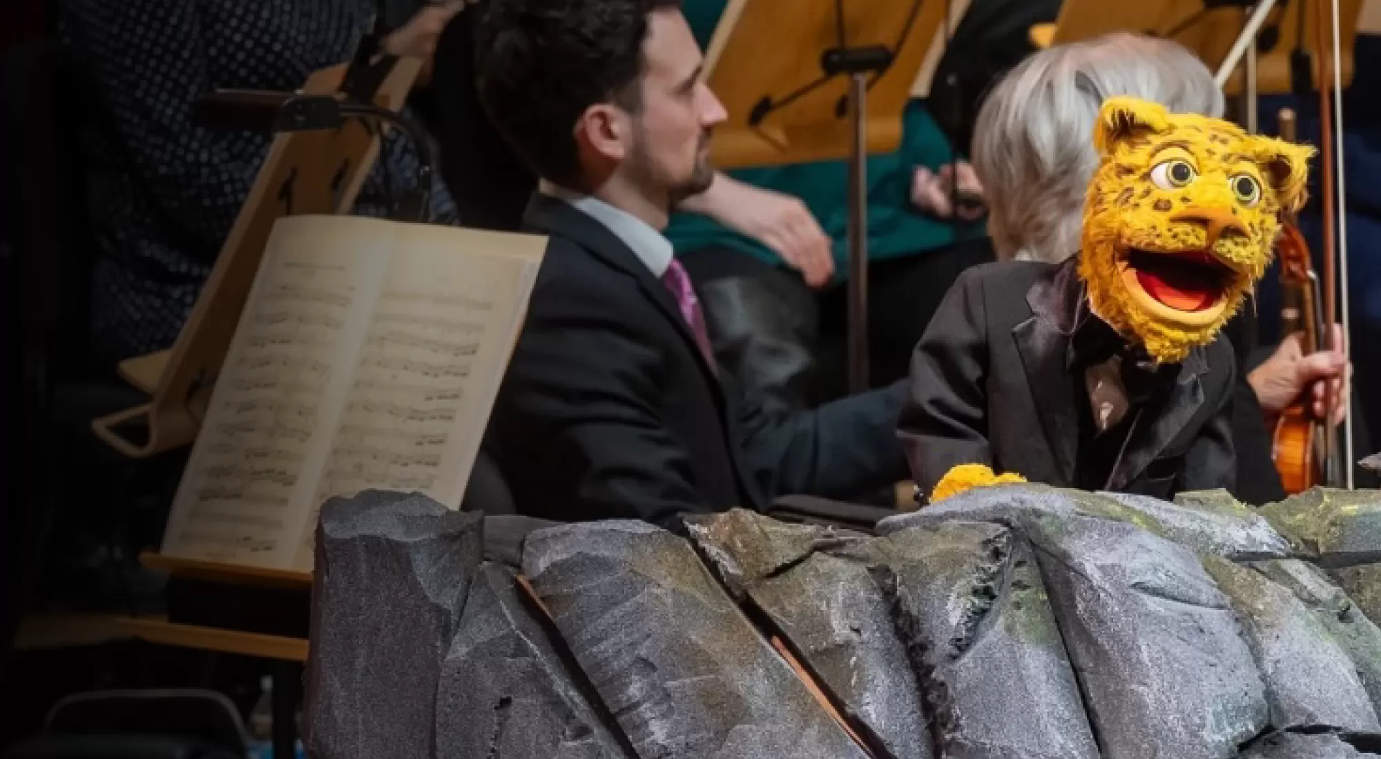 A furry leopard puppet on a rock in front of an orchestra