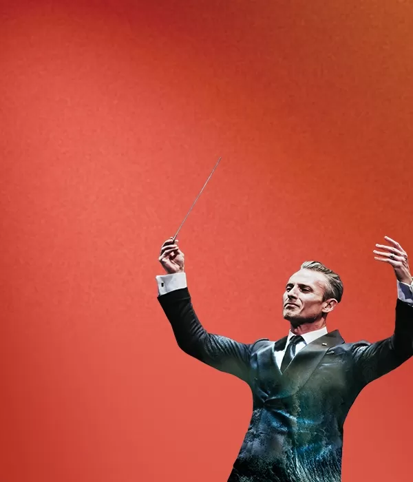 Alexander Shelley in a suit holding his baton in the air against a red background
