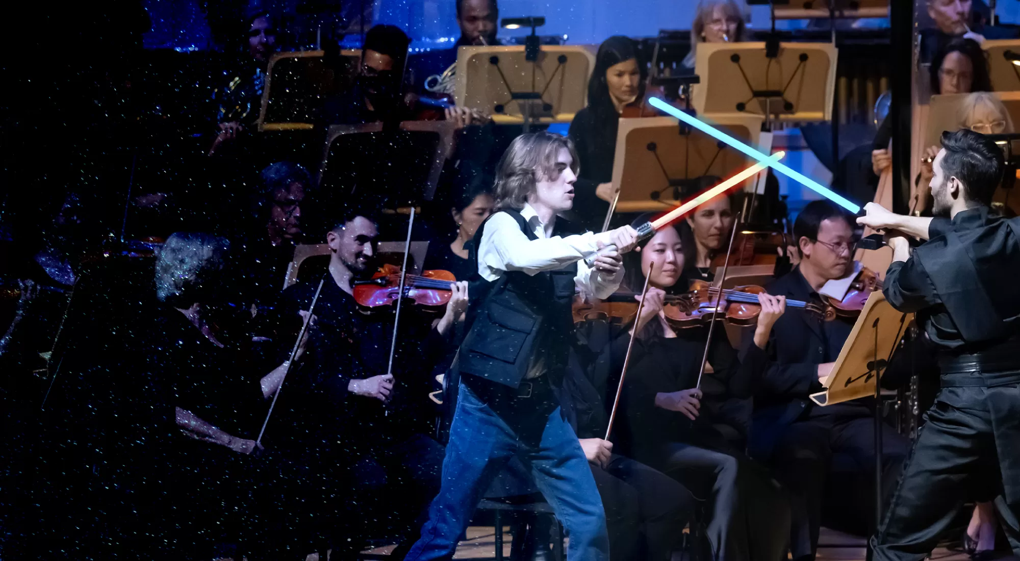 Two people have a light saber duel on stage in front of an orchestra