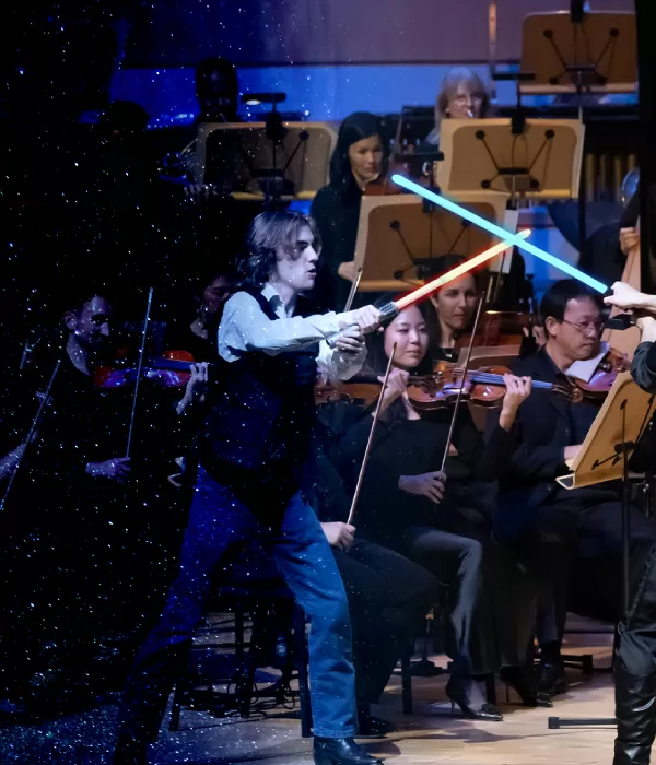 Two people have a light saber duel on stage in front of an orchestra