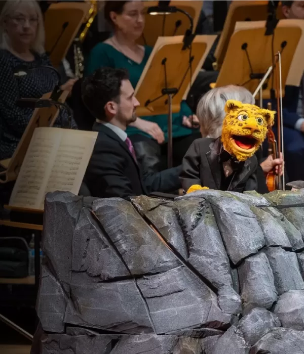 A furry leopard puppet on a rock in front of an orchestra